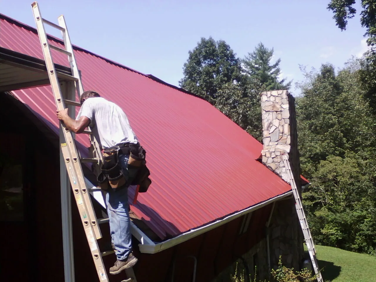 Roofer on a ladder beside a red metal roof and stone chimney — Soffit & Fascia Repair by Roofing Brothers in Sidman